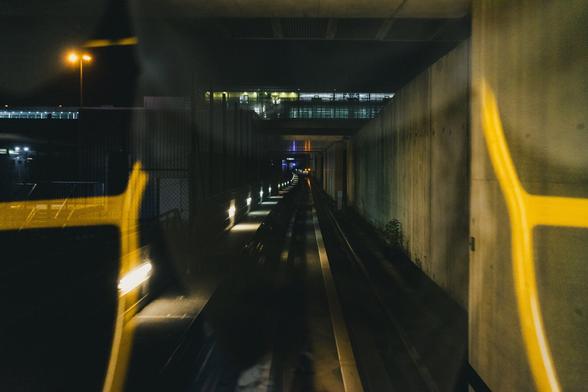The image shows a view from inside a transportation tunnel, likely from a train or tram looking down the tracks. It's nighttime, and the tunnel is dimly lit with lights along the track. There are reflections on the window, possibly from the inside of the vehicle. The scene extends into the distance, creating a sense of depth.