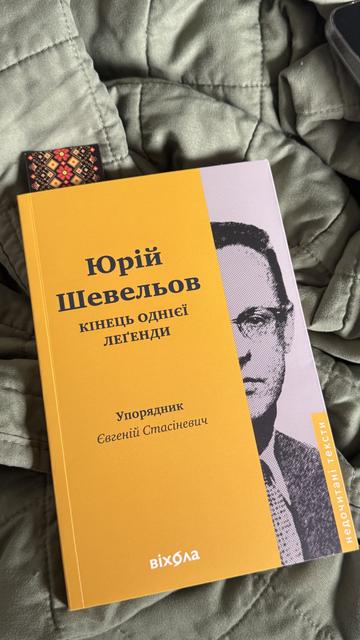 Ukrainian-language book titled Юрій Шевельов: Кінець однієї легенди (“Yuriy Shevelyov: The End of a Legend”), compiled by Євгеній Стасіневич (Yevhenii Stasinevych), lies on a soft, light-green quilted fabric. The book cover is mostly yellow with a partial black-and-white image of Shevelyov’s face. A woven Ukrainian-style bookmark with a red and black geometric pattern peeks out from the top.