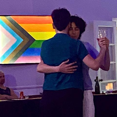 Two people dance with a glowing pride flag in the background. The back of a short-haired person in dark blue faces the camera. That person is following. Across their back, we see the leader’s arm, and over their shoulder, the curly-haired leader’s face.