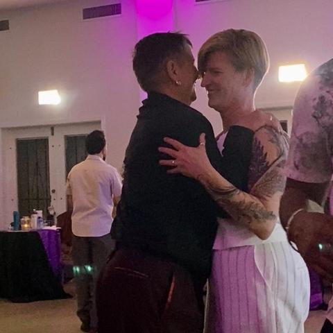 A pair of wives embrace with huge smiles of absolute joy. One is dressed in dark dress shirt and pants with very short hair and small hoop earrings. The other is dressed in a white waistcoat and white pinstriped pants.
