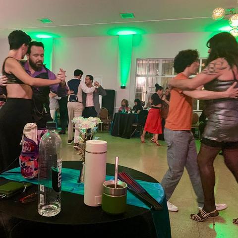 A scene of the dance floor. To the left, a man in purple leads a woman with short hair and a crop top. To the right, a shorter person in orange leads a taller woman in a silver dress. Behind, a pair of men dance.