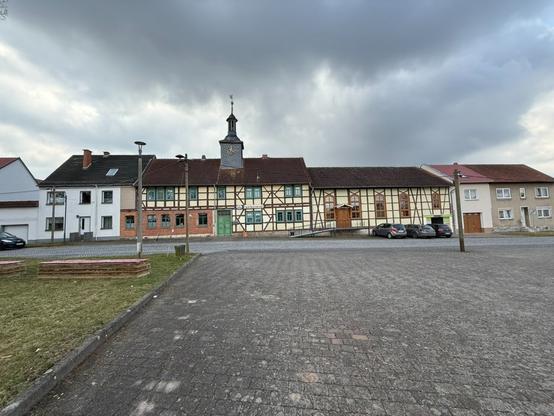 A street view featuring a series of traditional German half-timbered buildings alongside a gray sky. The scene includes a cobblestone area with parked cars, and a prominent clock tower on one of the buildings.