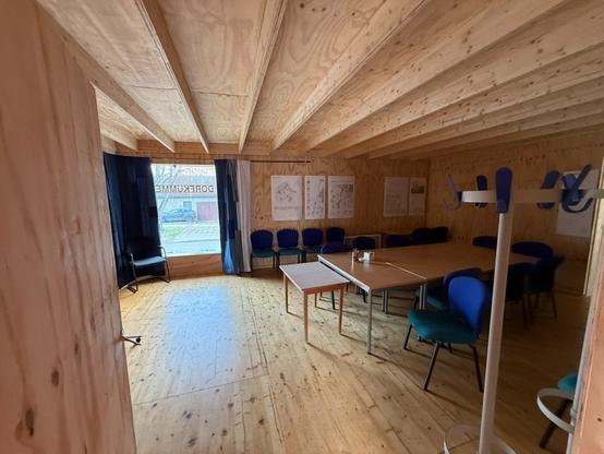 Interior of a wooden meeting room featuring a large table, several blue chairs, and large windows with dark curtains. One corner displays charts or plans on the wall. A coat rack is visible in the foreground.