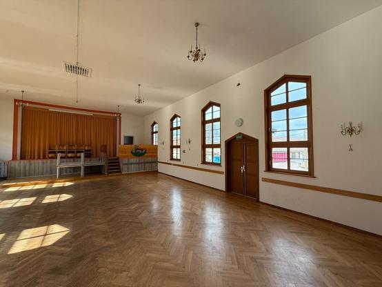 An empty hall with wooden parquet flooring, featuring large windows and a stage at one end with a curtain. There are a few chairs on the stage, and decorative wall sconces. Sunlight casts shadows on the floor.