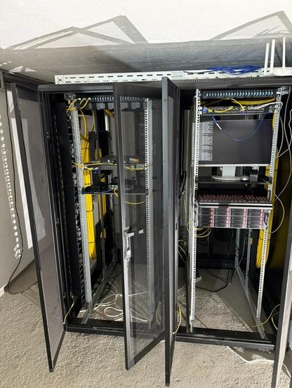 A server room containing two open metal rack cabinets. One cabinet holds networking equipment with visible cables, while the other features a computer monitor and additional equipment. The walls are bare, and the floor is carpeted.