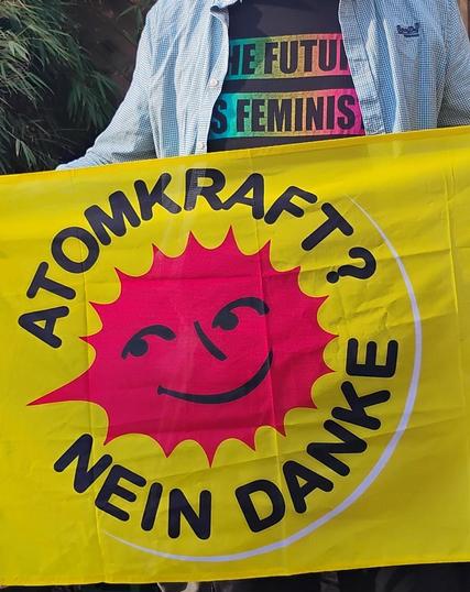 Flagge mit Roter Anti atom Kraft Sonne auf gelbem grund. Schwarzer schriftzug im Kreis um die Sonne: Atomkraft? nein danke. Ich halte die Flagge und Mensch sieht mein schwarzes T-Shirt und grünes offenes Hemd. Auf dem T-Shirt steht: the future is feminist.