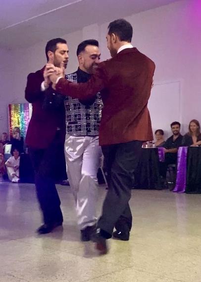 Twin brothers Nicolás &amp; Germán Filipeli dance with Leonardo Sardella between them. Germán is leading. All three men have short dark hair and beards. The twins are wearing maroon blazers and black pants. Leonardo is wearing a black shirt, black and silver plaid waistcoat, and silver pants. Nicolás looks serious. Leonardo is grinning.