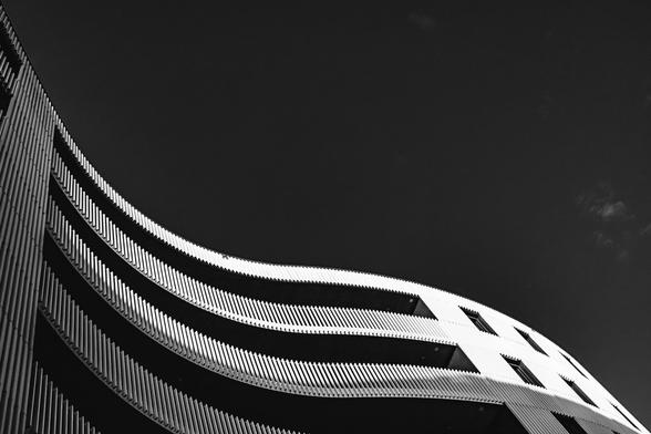 Black and white photo of a modern architectural building with wavy, horizontal lines and a rhythmic pattern of vertical slats. The curves of the structure contrast sharply against a clear, dark sky, giving the image a dynamic and minimalist feel.