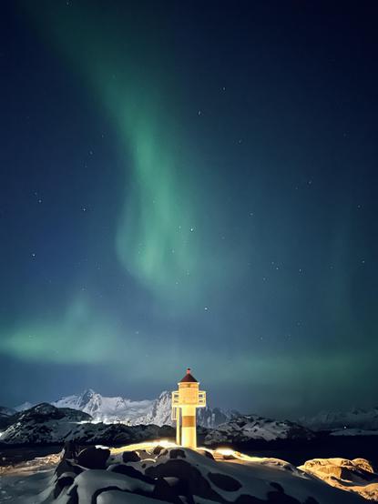 Grünes Polarlicht erhebt sich wie ein Lichtschweif am sternbedeckten Nachthimmel über einem beleuchteten Leuchtturm in einer verschneiten, bergigen Küstenlandschaft