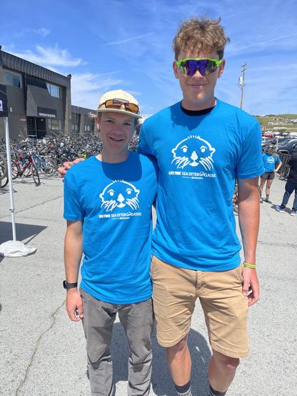 Two young friends wearing turquoise Sea Otter volunteer t-shirts stand together in the sunshine at the bicycle valet parking station. Many parked bicycles are visible in the background.