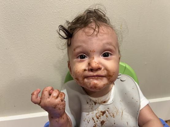 A light-skinned baby with curly hair sits in a highchair, covered in food, with puree smeared on their face and hands. They have a mischievous expression and one hand held up in a fist.