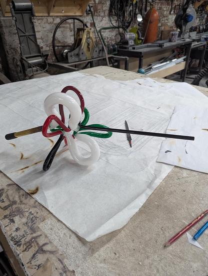 The forbidden doodle with side mismatches, a cut, a weld, and two bends away from completion. Big white loops, smaller green tube glass loops, smaller red tube loops, and even thinner black tube loop with another long straight black tube coming off. On a piece of charred paper, on drywall