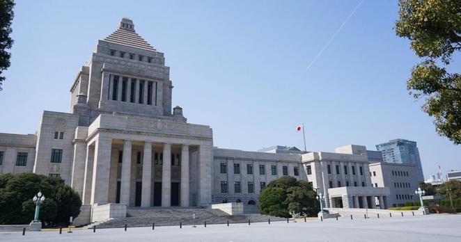 A large stone government building with a central pyramid-shaped tower, columns at the entrance, and a Japanese flag on a flagpole, surrounded by trees under a clear blue sky.