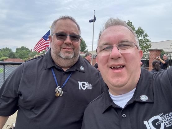 Two men smiling for a selfie outdoors, both wearing black polo shirts with logos. An American flag is in the background, along with people and trees. The scene appears to be part of an event or gathering.