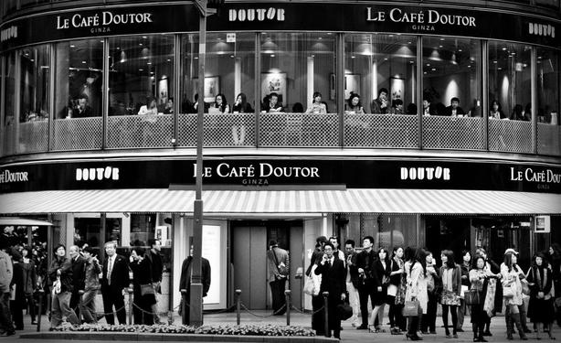 Vista de Le café doutor: un café de dos plantas. En la planta baja solo vemos personas paradas en la acera con aspecto de ir a trabajar (hay un hombre con maletín hablando por teléfono) o de compras. En la primera planta vemos una hilera de personas sentadas mirando a la calle aparentemente tomando café o comiendo algo, la mayoría parecen ir solos. Blanco y negro.