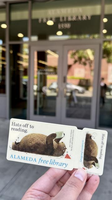 photo of an Alameda Free Library card featuring Jon Klassen’s bear (from I Want My Hat Back children’s book). The bear is lying on his back with his hat to the side, a book in his paws: he is reading Crime and Punishment. Next to him, there’s text: “Hats off to reading.” The photo is taken in front of the main branch of the Alameda Free Library.
