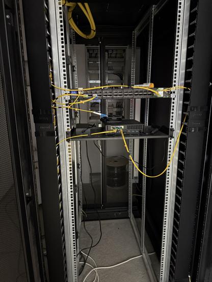 A server rack with patch panels and several yellow and black network cables. The interior features empty spaces for equipment, visible mesh panels, and a few mounted devices. The floor is gray with exposed wiring.