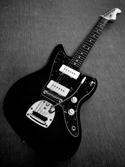 A black and white photo of a Jazzmaster electric guitar lying flat down on a plain carpet as seen at an angle from back end of the instrument.