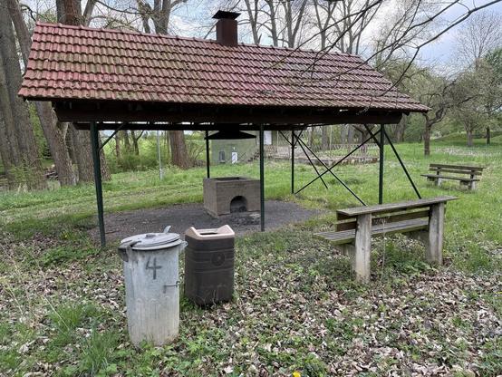 An outdoor picnic area featuring a covered grill with a red tiled roof, two trash bins, and benches. The surrounding area has grass and trees, creating a natural setting.