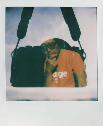 A Polaroid features a man wearing an orange shirt. He is sitting thoughtfully, partially obscured by a camera strap overhead. The background includes cloudy skies, giving the image a soft, dreamy quality