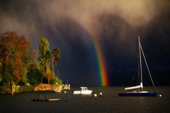 Dramatische Stimmung am Thunersee mit einem Regenbogen, der aus den Wolken direkt in den See zu stechen scheint. Links im Bild ist das Ufer mit Bäumen und einer Schiffsanlegestelle; auf dem See ein Badefloss und zwei Boote an Bojen.