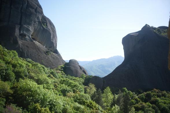 Two giant rocks (Meteora) are visible on the left and right. Below them towards the photographer are trees and vegetation. A mountain is visible between the two Meteora in the distance