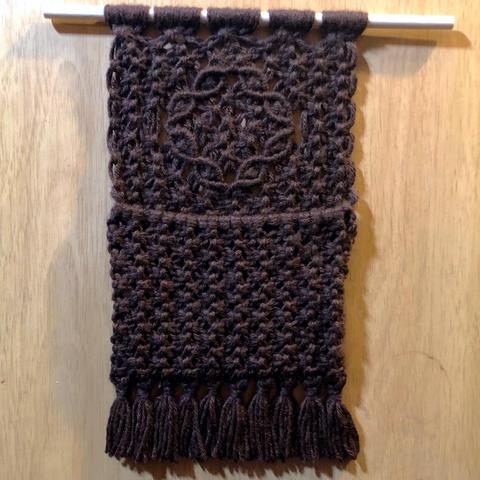 A dark brown  macrame hanging with a pouch at the bottom and tassels, all suspenden from a dowel. The pouch and the section above it are approximately the same height, but the pouch is entirely square knots, while the upper part has a circular flower like motif made of lines of half hitches arranged in petal like shapes with a grouping of square knots in the center.