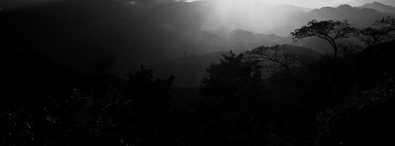 The image is a black and white landscape featuring a range of mountains. There are silhouettes of trees in the foreground, and the light is creating a dramatic effect with shadows and highlights across the scene. The overall mood is atmospheric and serene.