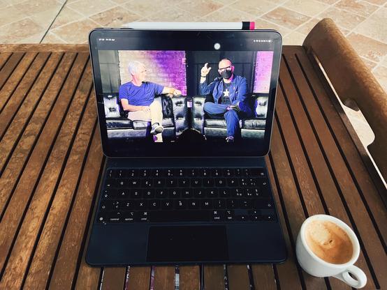 A photo of an iPad sitting on a table showing on screen a frame from Cory Doctorow’s conversation with Mike McCue at Fediverse House - they’re sitting on stage next to each other. Next to the iPad there is a small cup of coffee
