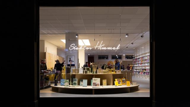 View through a large window into a brightly lit bookstore with a modern, minimalist design. A neon sign reading 'Siebter Himmel' (German for "Seventh Heaven") hangs from the ceiling. Inside, people browse books and decorative items displayed on round tables and tall shelves lining the walls.