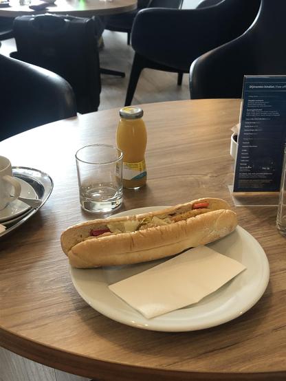 A quite huge sub on a plate, surrounded by pineapple juice and a cup of coffee. In thebackground, some stylish furniture lurking.