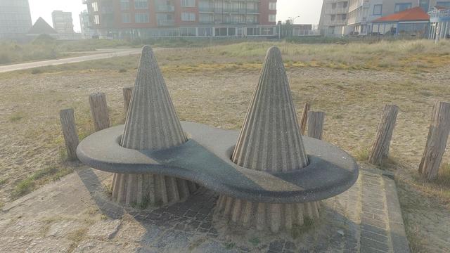 Public bench, with an 8 shaped seating area, and two cone shaped pyramids protruding through the two holes of the 8.