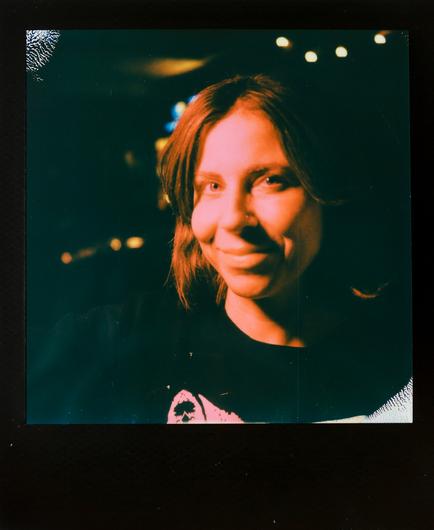 a black-framed Polaroid photo of a smiling woman illuminated in pinkish-red light with abstract lights from pinball machines in the background.
