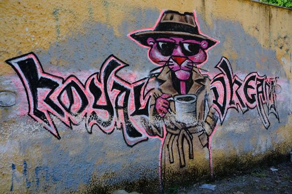 a graffiti on a yellow wall. the graffiti says “royal dream”, with a pink panther figure between the two words. the pink panther is holding a cup, wears sunglasses and a detective’s hat