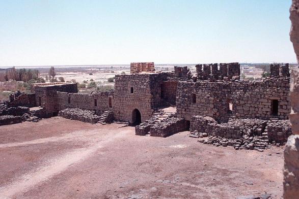 a photo of a stone castle complex, taken in a three quarter view, it is in the middle of the desert, some little shrubs and trees around it, a bright blue sky overhead