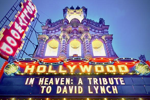 The front facade of a palatial-esque movie theatre bears a giant vertical sign that reads, "HOLLYWOOD" with white lettering over a red background, and a marquee below it that reads, "IN HEAVEN: A TRIBUTE TO DAVID LYNCH" in gold/white lettering over a dark blue background, all adorned with bright lights around the borders under a blue evening sky.