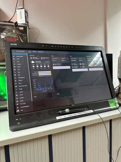 Ein 24-Zoll großes Touchdisplay, auf dem unser Home-Assistant Interface gezeigt wird. Anzeigt wird ein Dashboard, welche den Stromverbrauch über die Zeit, das Wetter den Space-Status, die jeweilige Temperatur und CO2-Werte unserer Räume beinhaltet sowie, dass auf den Chromecasts derzeit nichts läuft. Links werden weitere Dashboards aufgelistet wie etwa für die Räume oder für den Strom. Unter den Touchpanel ist Stift für diesen, an den mit Heißkleber ein Knopf gepfuscht wurde. Daruter ein Labelprinter-Hinweis: "Stift mit ausreichend Druck aufdrücken". Unten rechts klebt ein Public-Sticker auf den der Name "Andi" als Besitzer geschrieben wurde. Unten links ist der Powerbutton. Das Display steht auf einem Regal und ist von diverser Elektronik umzingelt.