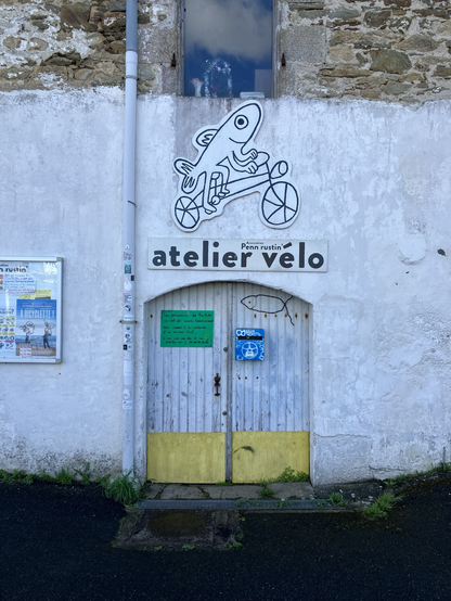 Devanture d’un atelier vélo. L’enseigne représente un poisson pédalant sur un grand vélo, dessiné à la main.