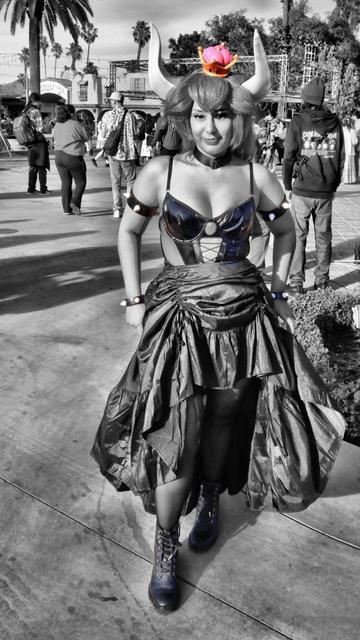 Woman in a cosplay as Bowsette.  Large white horns coming out of a red hair wig  with a little pink crown.  Arms are up touching the horns with spiked bracelets and arm bands.  A sexy corset and with sheer side and black dress and black boots.