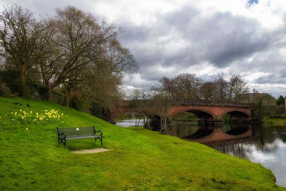 Frühlingsbild von Callander in Schottland mit einer Brücke über den Fluss 
Teith. Links im Bild ist eine unbesetzte Ruhebank auf einer Wiese mit Narzissen und Krähen.