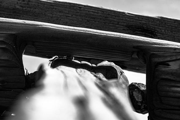 A groyne on Goring beach, a black and white image focussed on the wooden texture of the groyne, following it from the bottom of the image away from the viewer towards the groyne itself which forms an angled horizon across the sky. A shallow depth of field forces the focus into the joint of the timbers and the shadow that lies within.