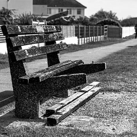 A bench facing Goring beach, framed such that the top of the bench falls on the leading line of the fences that separate the houses behind from the sea path. Two slats have fallen from the bench seat and been placed underneath. The black and white photo hints at an unfocussed figure just walking right, out of frame, the bench foregrounded to the left leading diagonally right towards the presence.