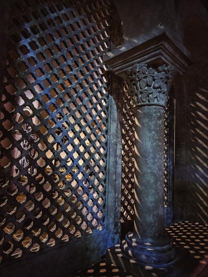 Detailed shot of a stone column with carved floral designs next to a dark, patterned lattice wall, with sunlight creating dappled shadows on the column and the floor.