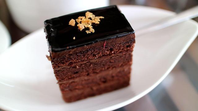 4 layer chocolate cake topped with nuts and chocolate ganache on a white, leaf shaped platter (background is blurred)