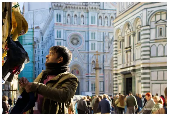 The picture shows a merchant arranging hats on the outdoor shop display. There is the Cathedral of Santa Maria del Fiore, and part of the Baptistery of St. John in the background.