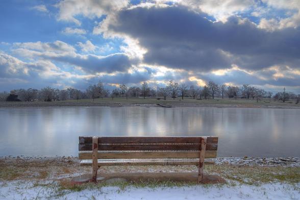 A wooden bench overlooks a tranquil lake, surrounded by snowy grass and under a dramatic sky filled with clouds.