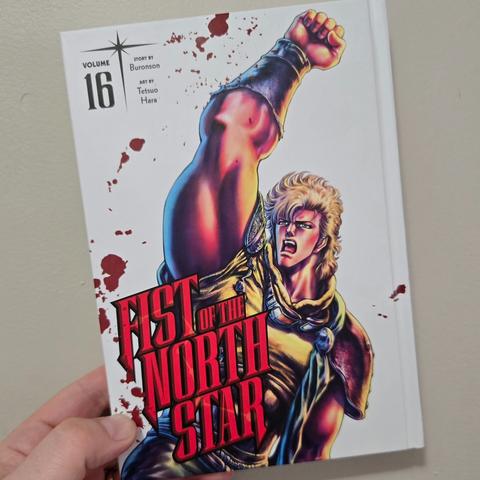 Photograph of a white person's hand holding Fist of the North Star, Volume 16, against a light colored wall. The hardcover book has a white cover with the number 16 in black text in the upper left-hand corner and "Fist of the North Star" in all red, uppercase lettering in the lower left-hand corner. A beefy, blonde man with his fist raised is on the cover behind the text. Blood streaks are strewn about the cover seemingly randomly.