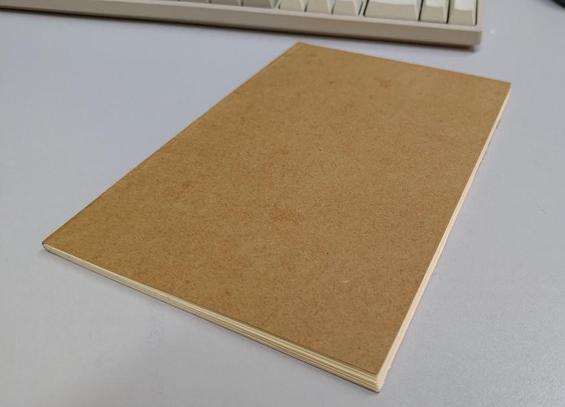 Foto de um caderno fechado sobre a mesa. A mesa é de fórmica cinza e o caderno é do formato A5 com capa de papel kraft em branco, com as folhas do miolo na cor creme. A lombada é simples, formada pelo papel Kraft da capa, sem espiral nem nada do tipo.

Ao fundo, fora de foco, dá pra ver um teclado mecânico bege retrô <3