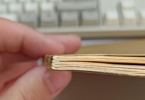 Detalhe da encadernação, com o caderno fechado, mostrando a lombada por baixo. Os quatro blocos dobrados ficam evidentes, com a capa de papel Kraft contornando a lombada como uma peça inteiriça.

Ao fundo, fora de foco, o teclado bege retrô <3