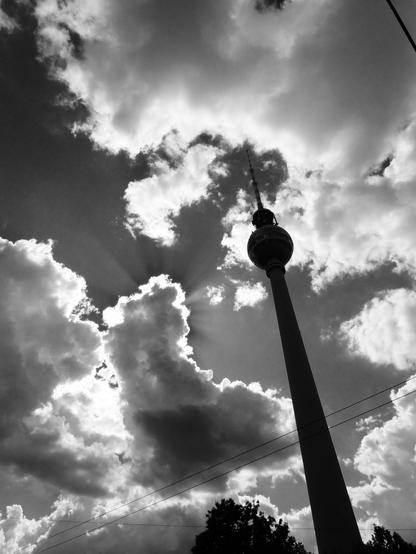 Black and white photograph of the Berlin TV Tower (Fernsehturm) silhouetted against a dramatic sky. The tower rises prominently from the bottom right of the frame, its distinctive spherical observation deck and antenna spire clearly visible. The sky is filled with billowing clouds. Sunbeams pierce through gaps in the clouds, radiating outward. In the lower portion of the image, power lines stretch horizontally across the frame, and the silhouettes of trees are barely visible at the bottom edge. The composition emphasizes the tower's height and the sky's turbulent nature.